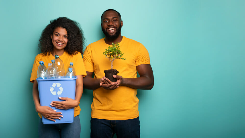 Happy couple hold a plastic container and a small tree over a light blue color. Concept of forestation, ecology, conservation, recycling and sustainability